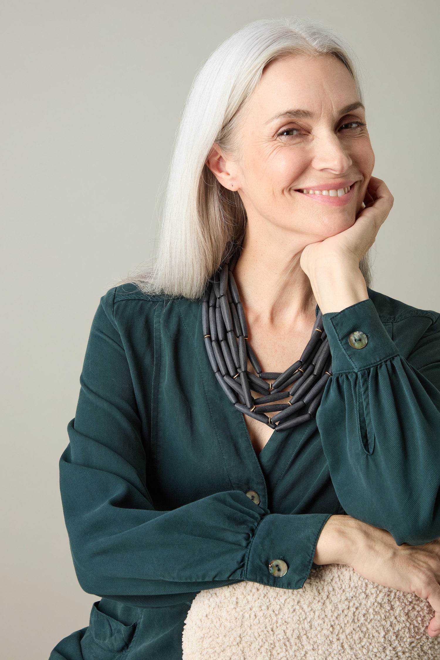 A woman with long gray hair, dressed in a green blouse, smiles as she rests her chin on her hand while wearing the exquisite Noir Cascade Necklace adorned with matte black beads.