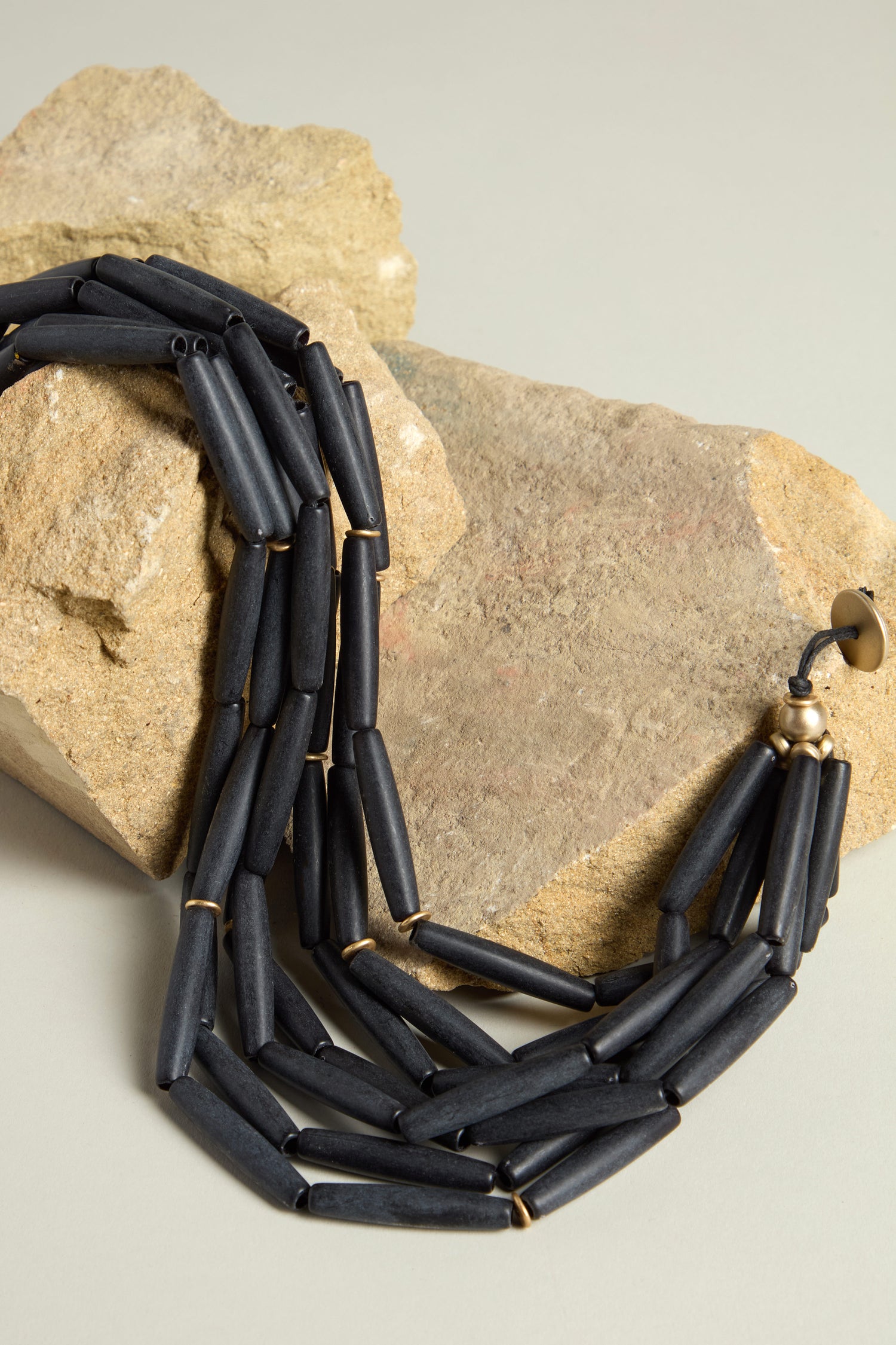 The Noir Cascade Necklace showcases a sleek aesthetic with multiple strands of matte black beads, gracefully arranged over a textured beige rock on a light gray surface.