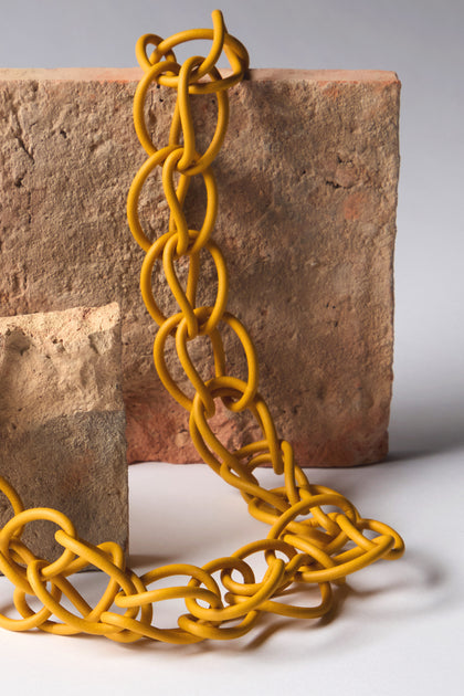 The Mustard Long Links Necklace, a modern accessory, showcases a twisted yellow chain crafted from flexible material. It elegantly complements two rectangular reddish-brown textured stones on a flawless white surface.