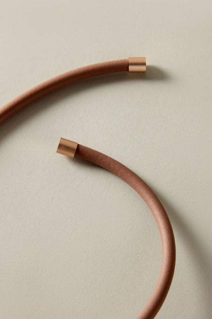 Against a light beige background, the Single Sphere Necklace showcases two brown, flexible cables with copper ends, embodying a minimalist and contemporary design.