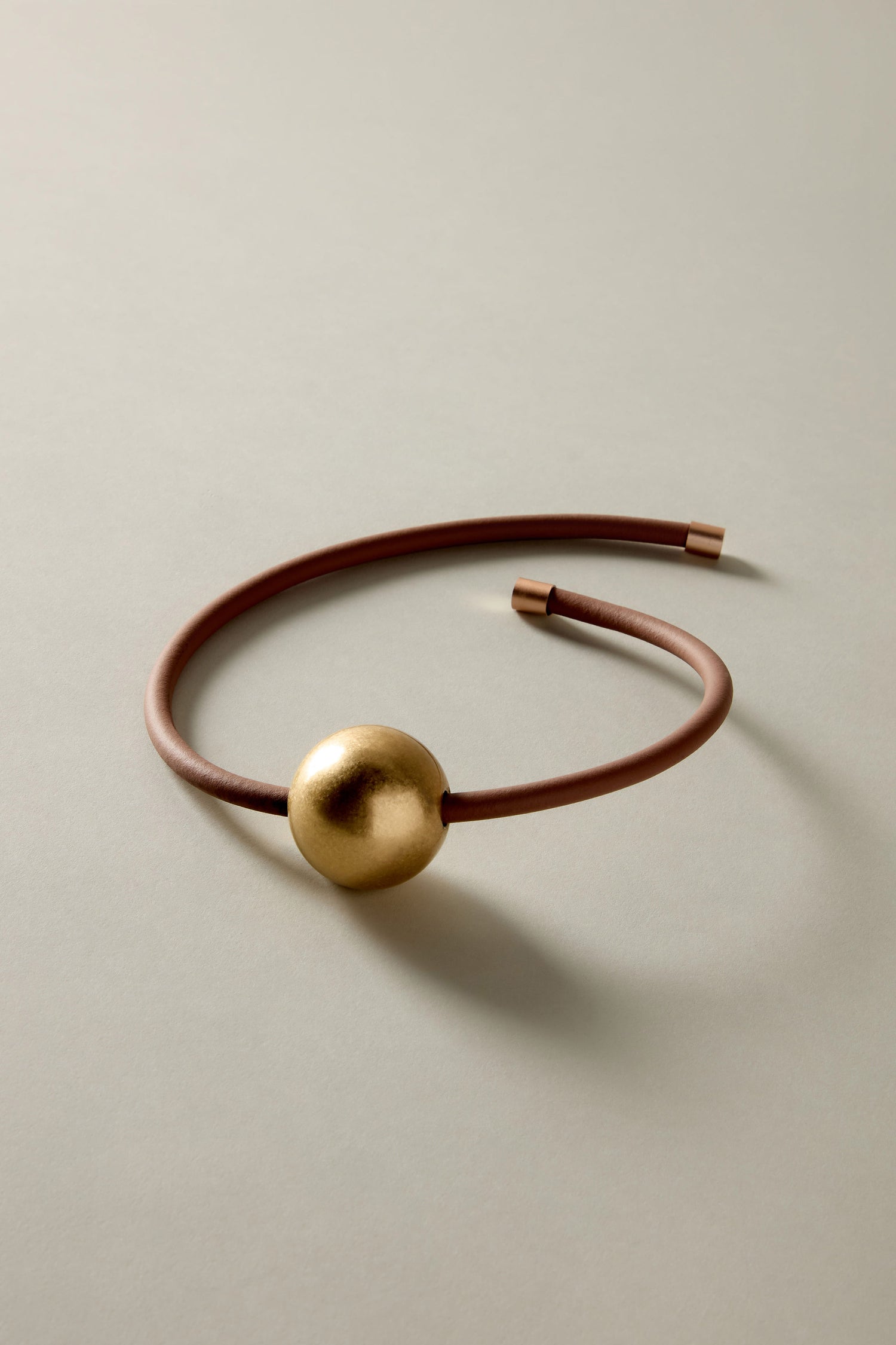 The Single Sphere Necklace is a brown leather choker with a large gold spherical bead, elegantly displayed on a light grey surface that highlights its minimalist, contemporary design.