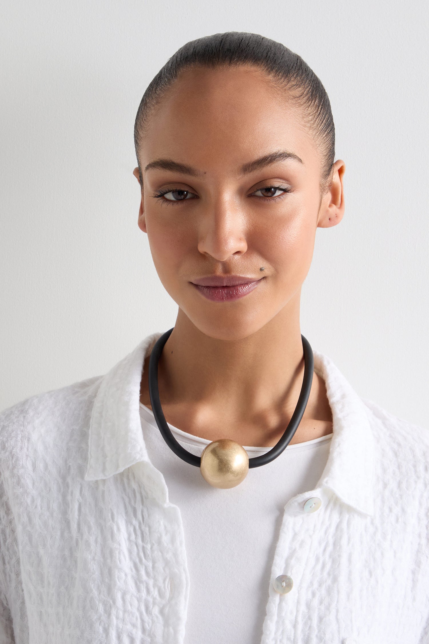 Wearing a white textured shirt and the Single Sphere Necklace, featuring a large gold orb, they embrace minimalist style while facing forward against a plain backdrop, exuding contemporary design.