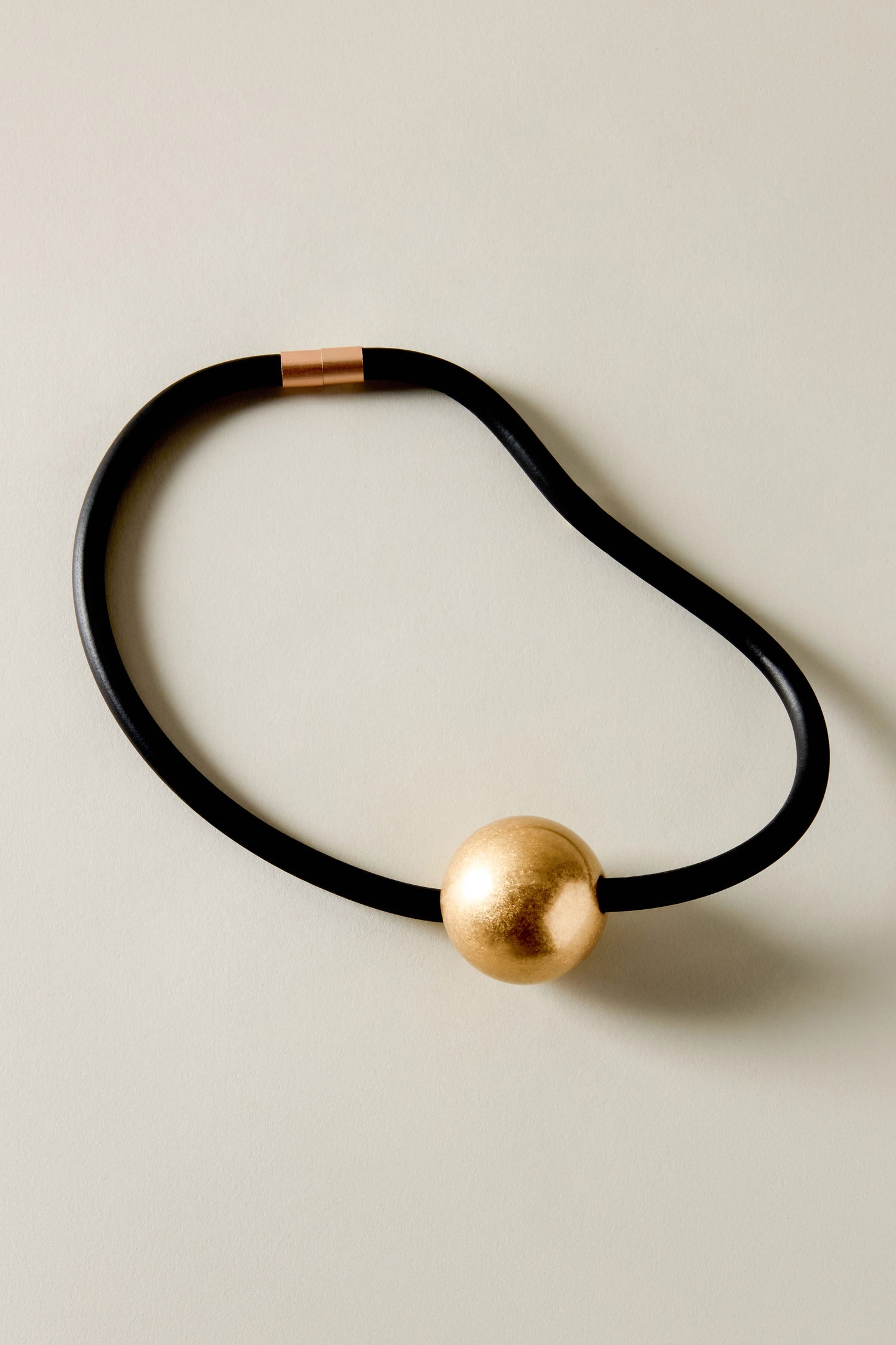 The Single Sphere Necklace showcases minimalist design, featuring a black cord with a large gold sphere pendant and a small gold clasp, elegantly resting on light surfaces.