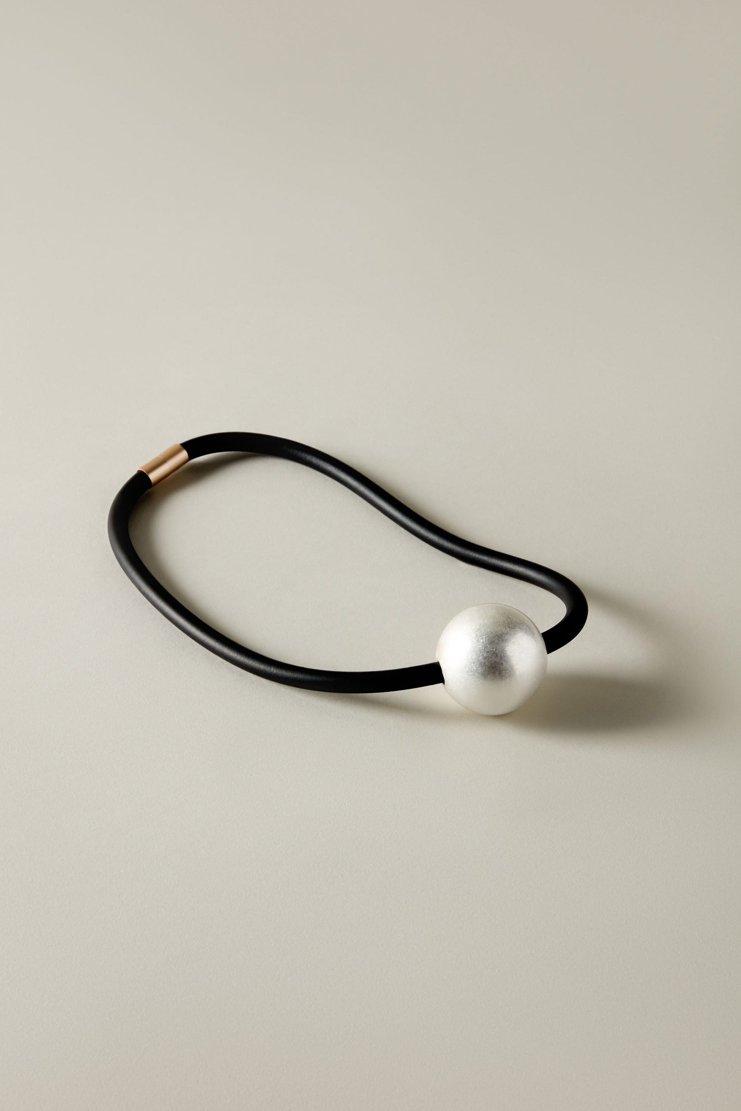 A black elastic hair tie with a large silver bead rests on a neutral background, showcasing the minimalist and contemporary design of the Single Sphere Necklace.