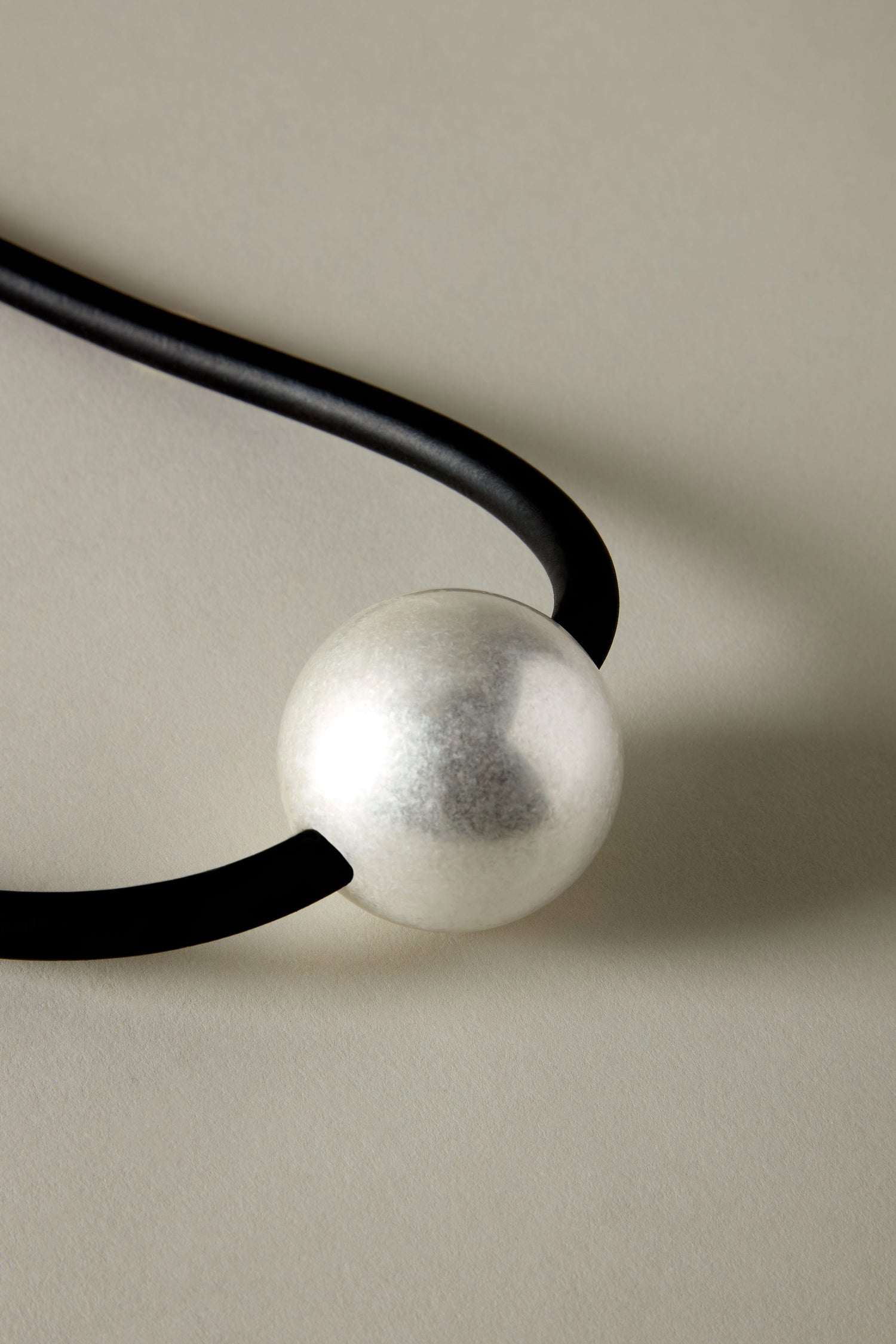 The Single Sphere Necklace showcases a minimalist design with its silver bead on a black cord, creating a contemporary and stylish accessory.