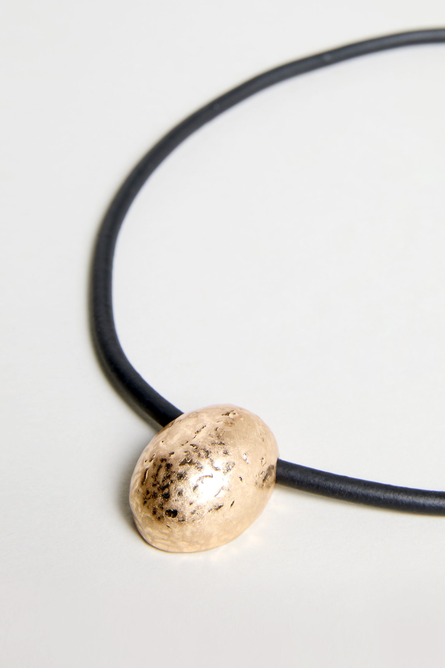 The Hammered Pebble Necklace features a black rubber cord and a singular oval hammered metal pendant, displayed on a light background.