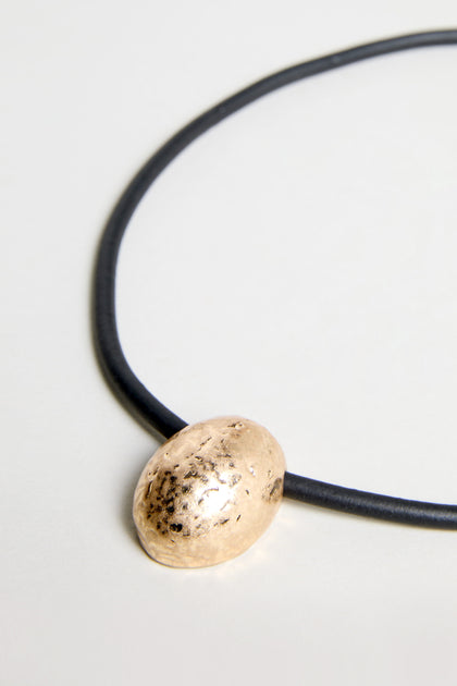The Hammered Pebble Necklace features a black rubber cord and a singular oval hammered metal pendant, displayed on a light background.