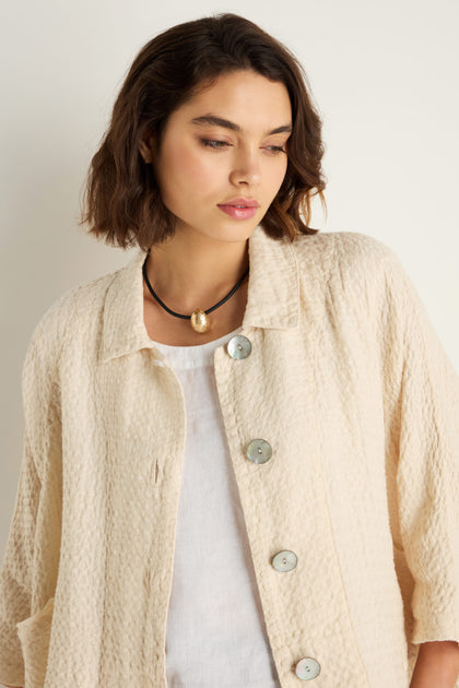 A woman with short brown hair stands against a plain background, wearing a textured cream button-up jacket over a white top and the Hammered Pebble Necklace, which features a gold pendant.