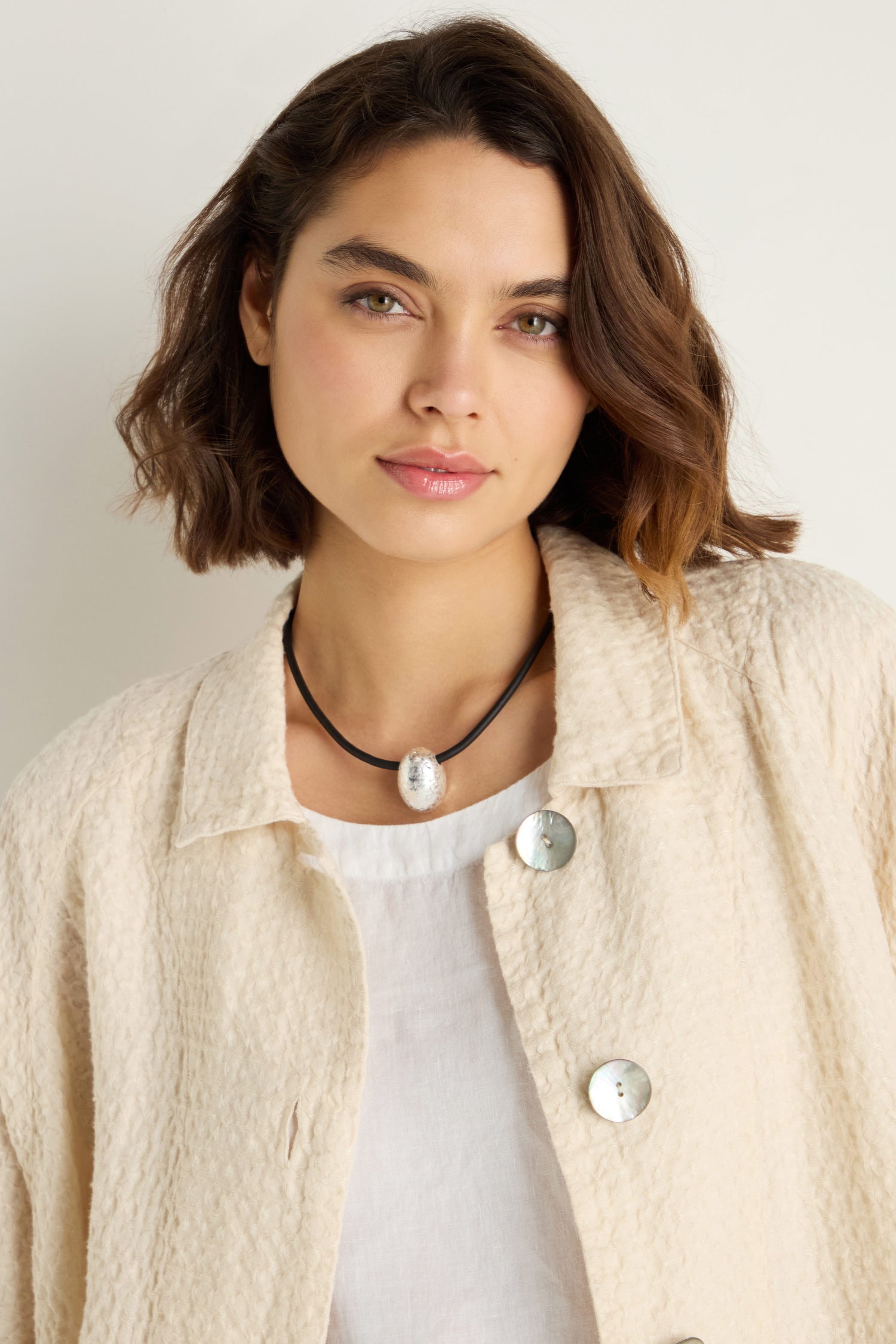 Woman with wavy brown hair wears a textured cream jacket, white top, and the Hammered Pebble Necklace—a hammered metal pendant on a black rubber cord—standing against a plain light background.