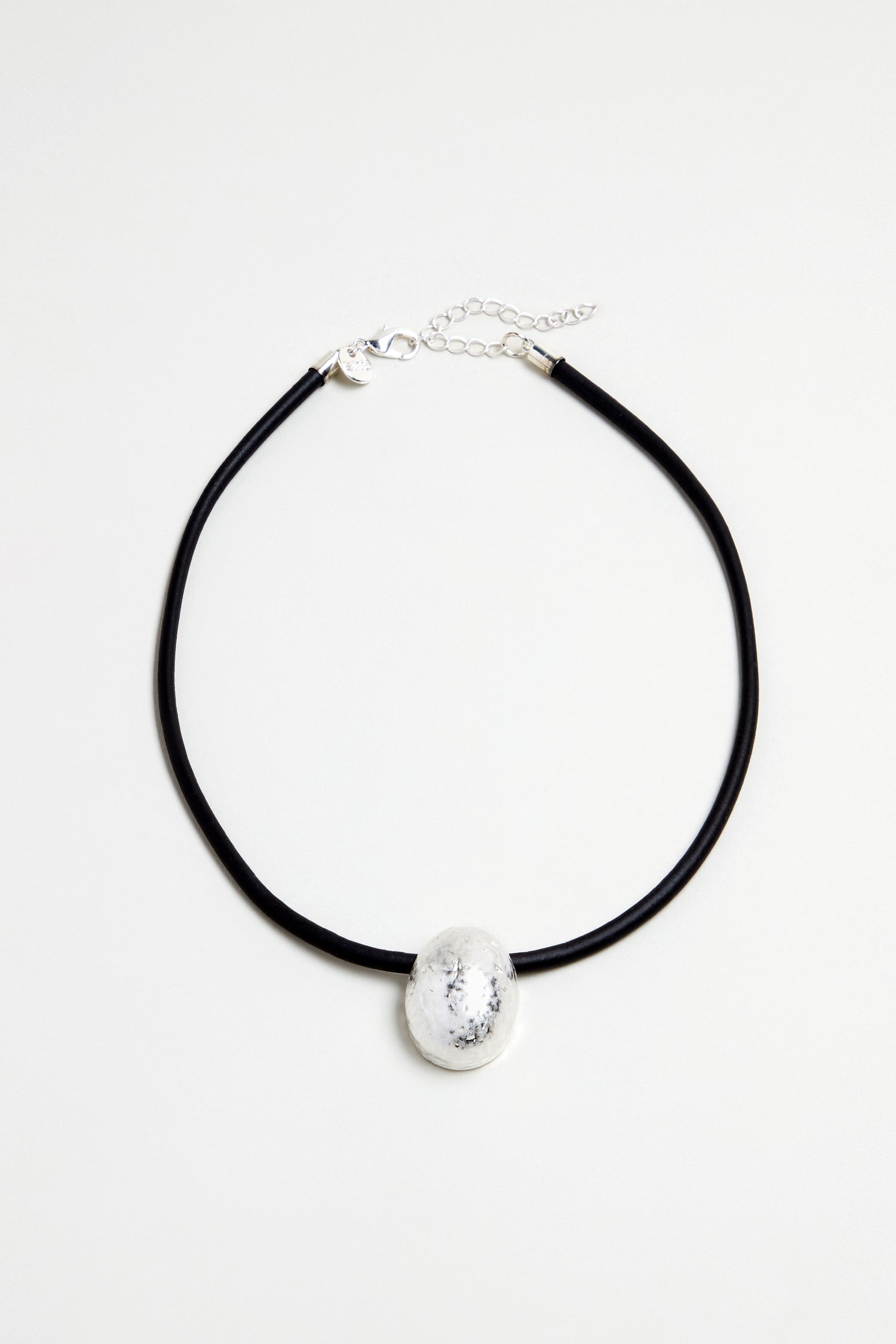 The Hammered Pebble Necklace features a black rubber cord with a large, round hammered metal pendant and clasp closure, shown on a plain white background.