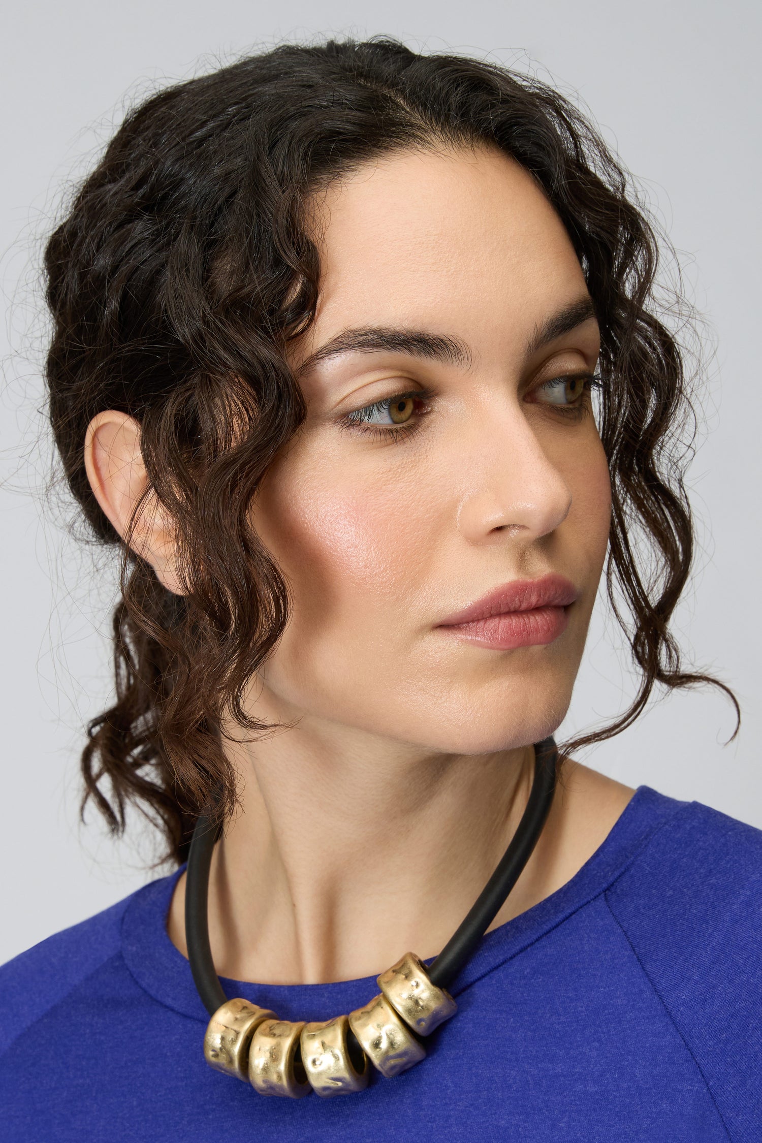 Woman with wavy dark hair wearing a blue top and the Hammered Cylinders Necklace, looking to her left against a plain background.
