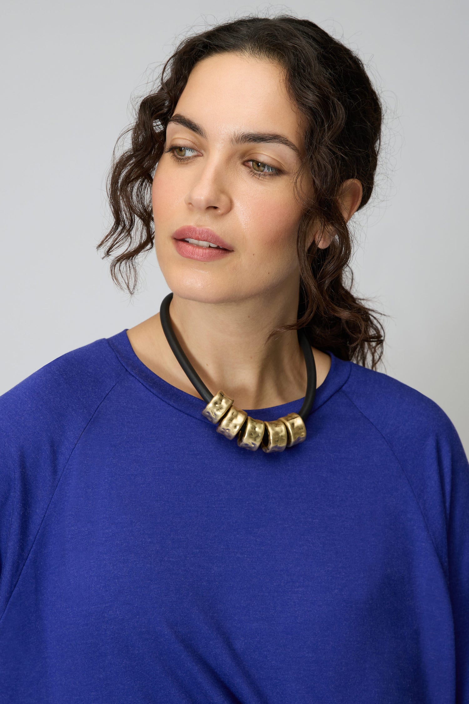 A woman with wavy brown hair in a blue top wears the Hammered Cylinders Necklace, featuring gold beads, as she gazes slightly to the side against a plain background.