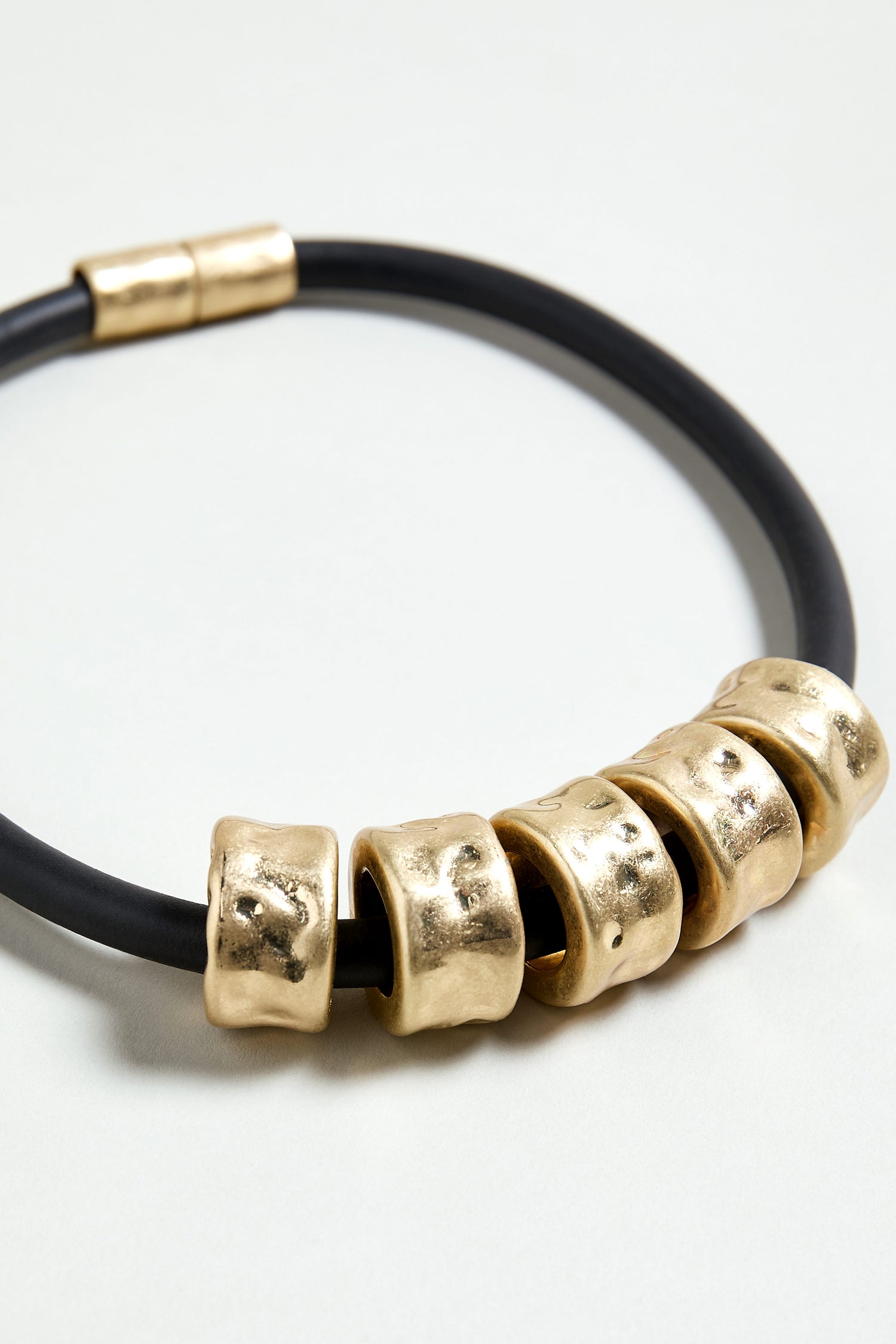 The Hammered Cylinders Necklace features five large, textured gold beads on a black cord, a sculptural contemporary design with a gold magnetic clasp. Shown on a white background.