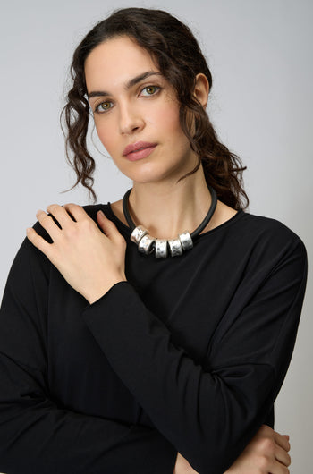 Woman with wavy dark hair wearing a black long-sleeve top and the Hammered Cylinders Necklace, posing with one hand on her shoulder against a plain background.