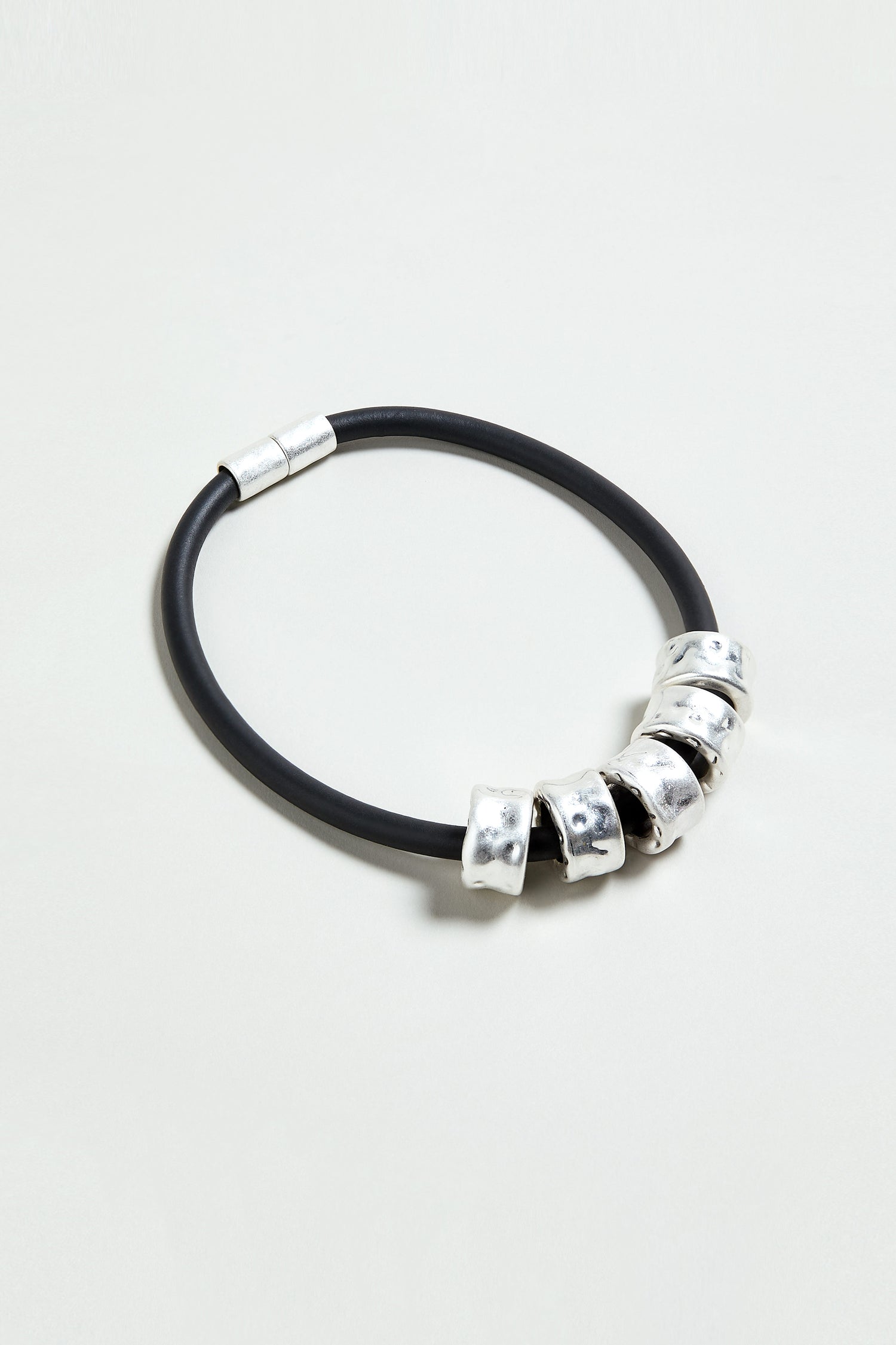 The Hammered Cylinders Necklace features five irregular silver beads on a black cord with a silver magnetic clasp, capturing contemporary elegance and style, all showcased against a light backdrop.