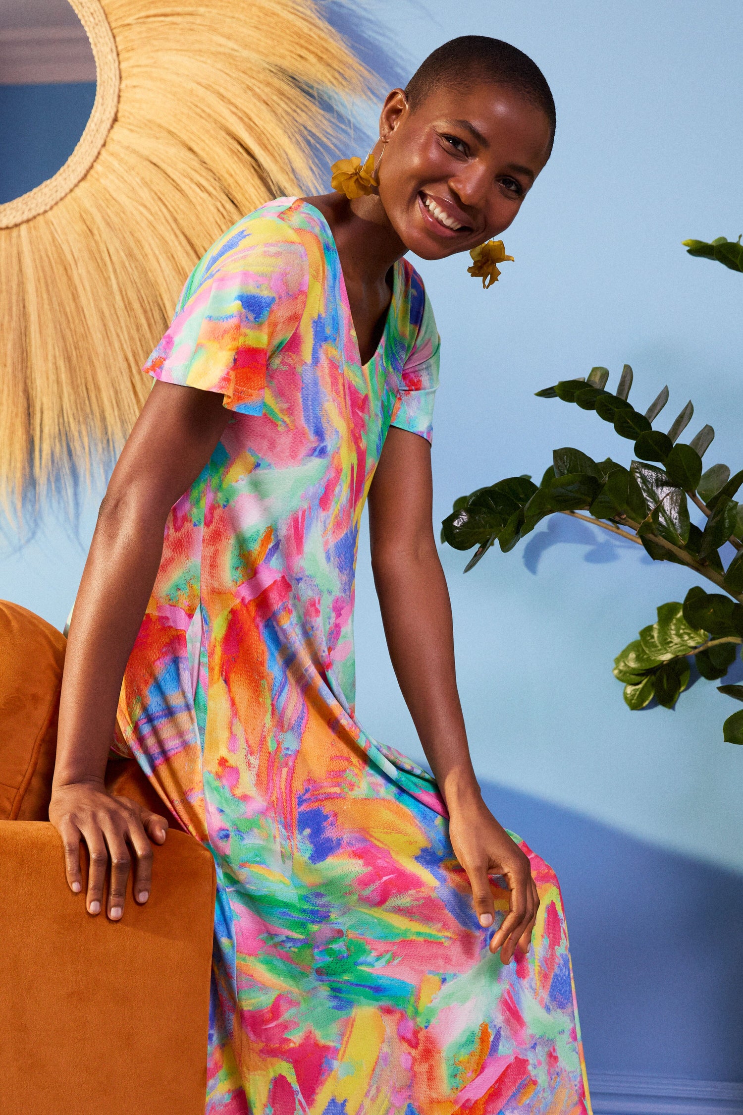 A person smiles while sitting on an orange sofa, donning a Vibrant Watercolour Jersey Dress and yellow earrings. Behind them is a blue wall with a sunburst mirror and a green plant.