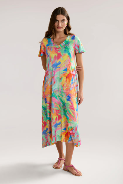 A woman is wearing a colorful, abstract-patterned Vibrant Watercolour Jersey Dress that reaches mid-calf. The LENZING™ Viscose ECOVERO™ fabric drapes elegantly on her relaxed figure, with one hand resting on her hip and her long hair cascading down.