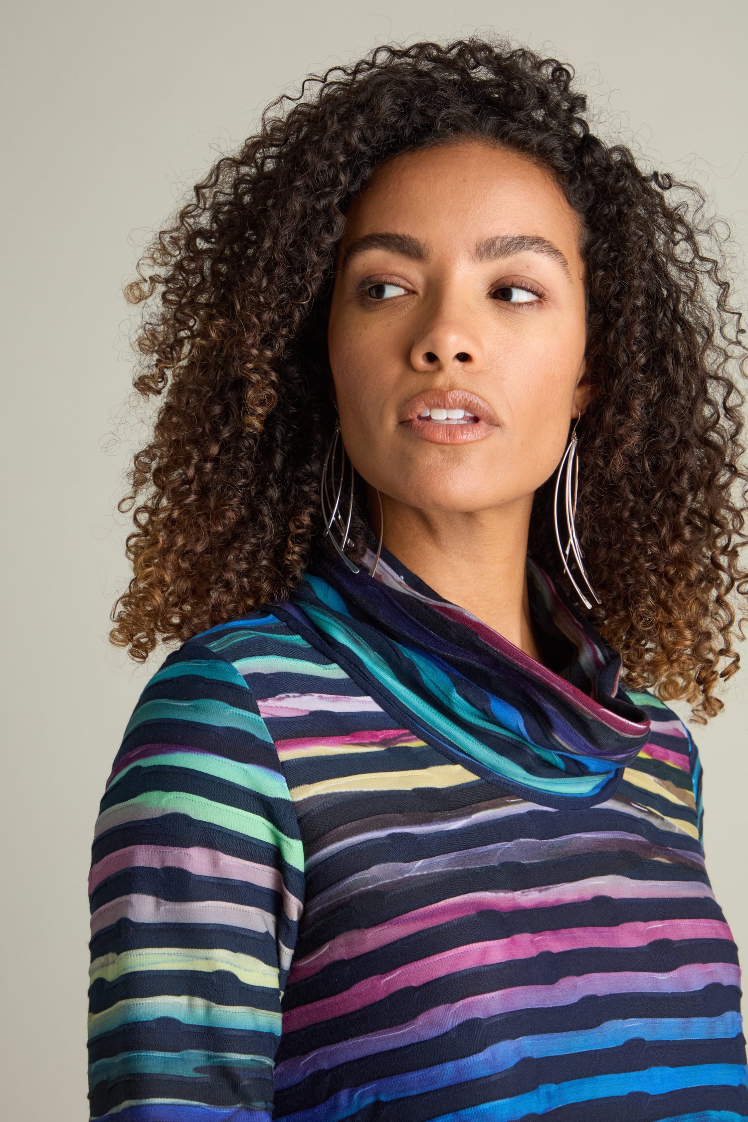 A woman with curly hair wears large hoop earrings and a Textured Watercolour Landscape Cowl Dress, looking slightly to her left.