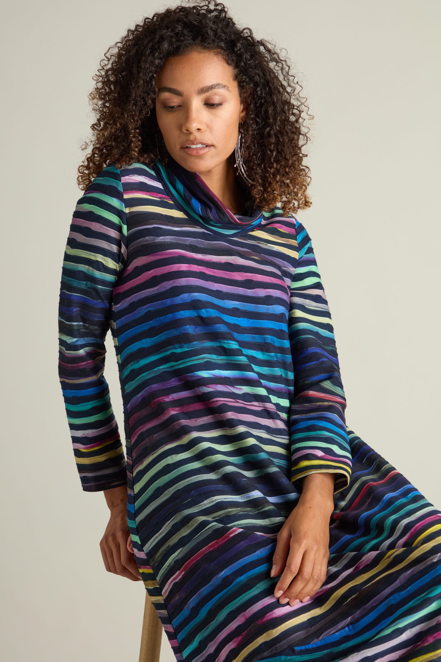 A woman with curly hair is seated, wearing the Textured Watercolour Landscape Cowl Dress, which features long sleeves and striking horizontal multicolored stripes in a beautiful ocean-hued watercolor print.