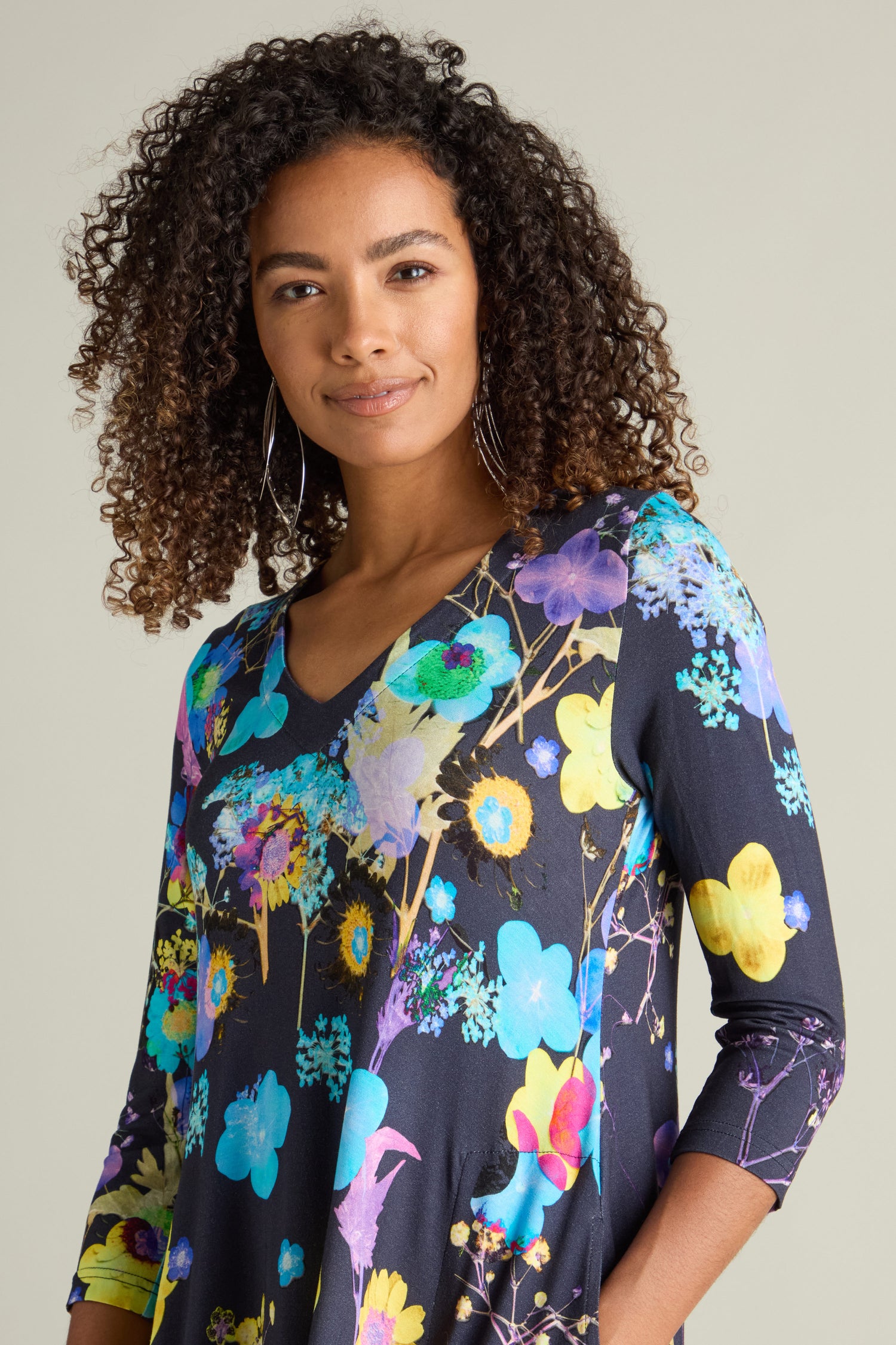 A person with curly hair stands against a plain background, showcasing the Blue Bouquet Jersey Dress adorned with three-quarter sleeves and a botanical print.