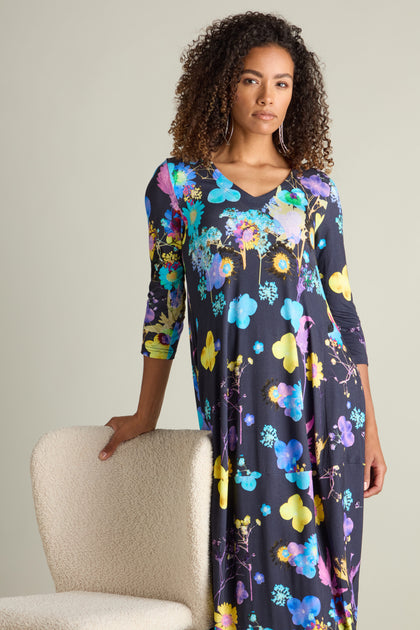 A person with curly hair is wearing a Blue Bouquet Jersey dress featuring a colorful botanical print and long sleeves, standing with one hand resting on a white chair.