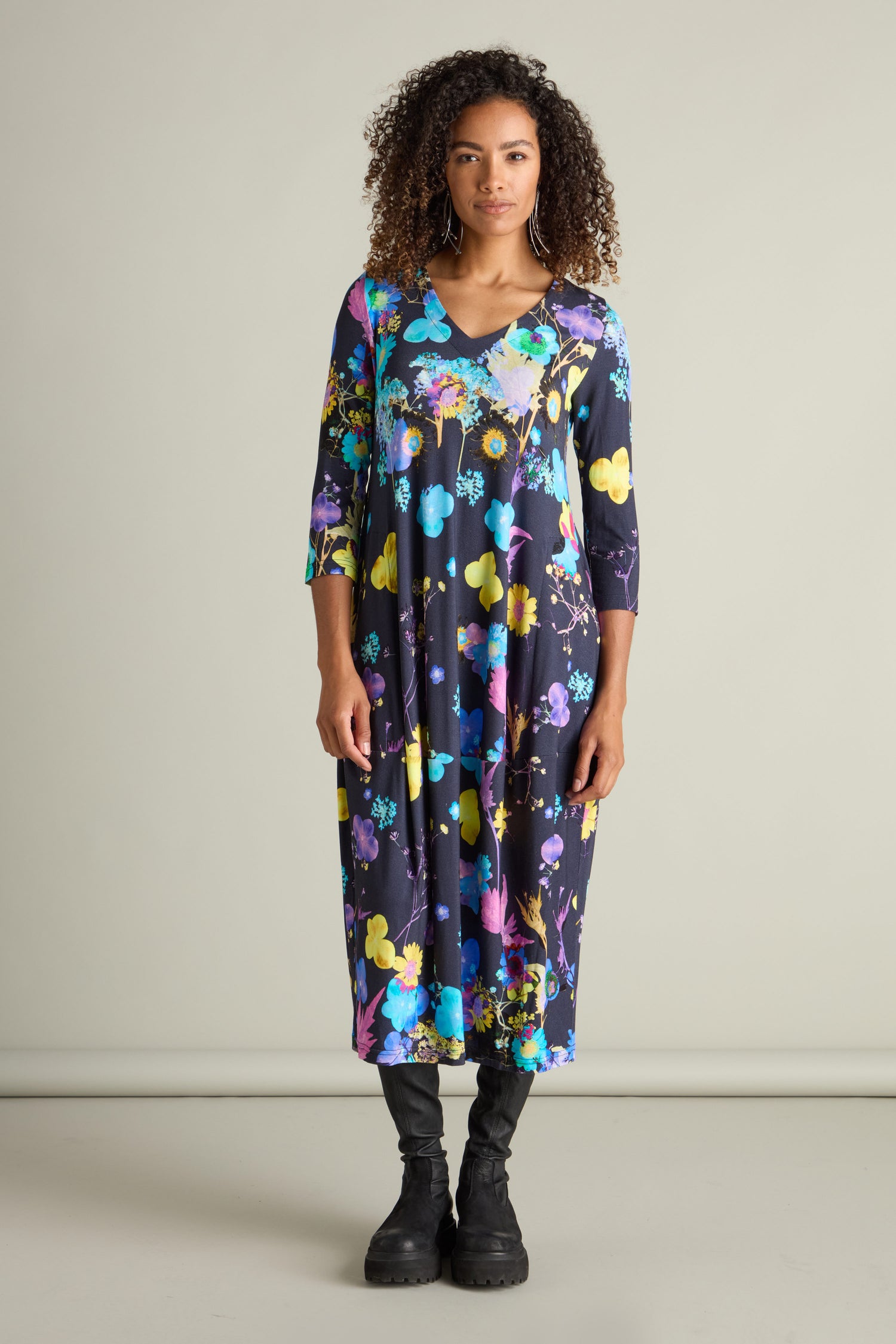 A woman stands facing forward, wearing a long Blue Bouquet Jersey Dress adorned with a colorful botanical print, paired with black boots. She has curly hair and a neutral expression.