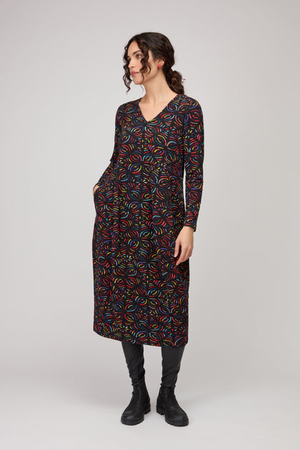 A woman wears the Mosaic Jacquard Jersey Bubble Dress—black, long-sleeved, knee-length with a colorful circular pattern—paired with black leggings and boots against a plain background.