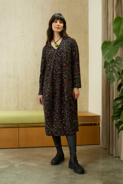 A woman stands indoors wearing a Mosaic Jacquard Jersey Bubble Dress with black boots and a neutral expression. Nearby are a green cushioned bench and a large plant.