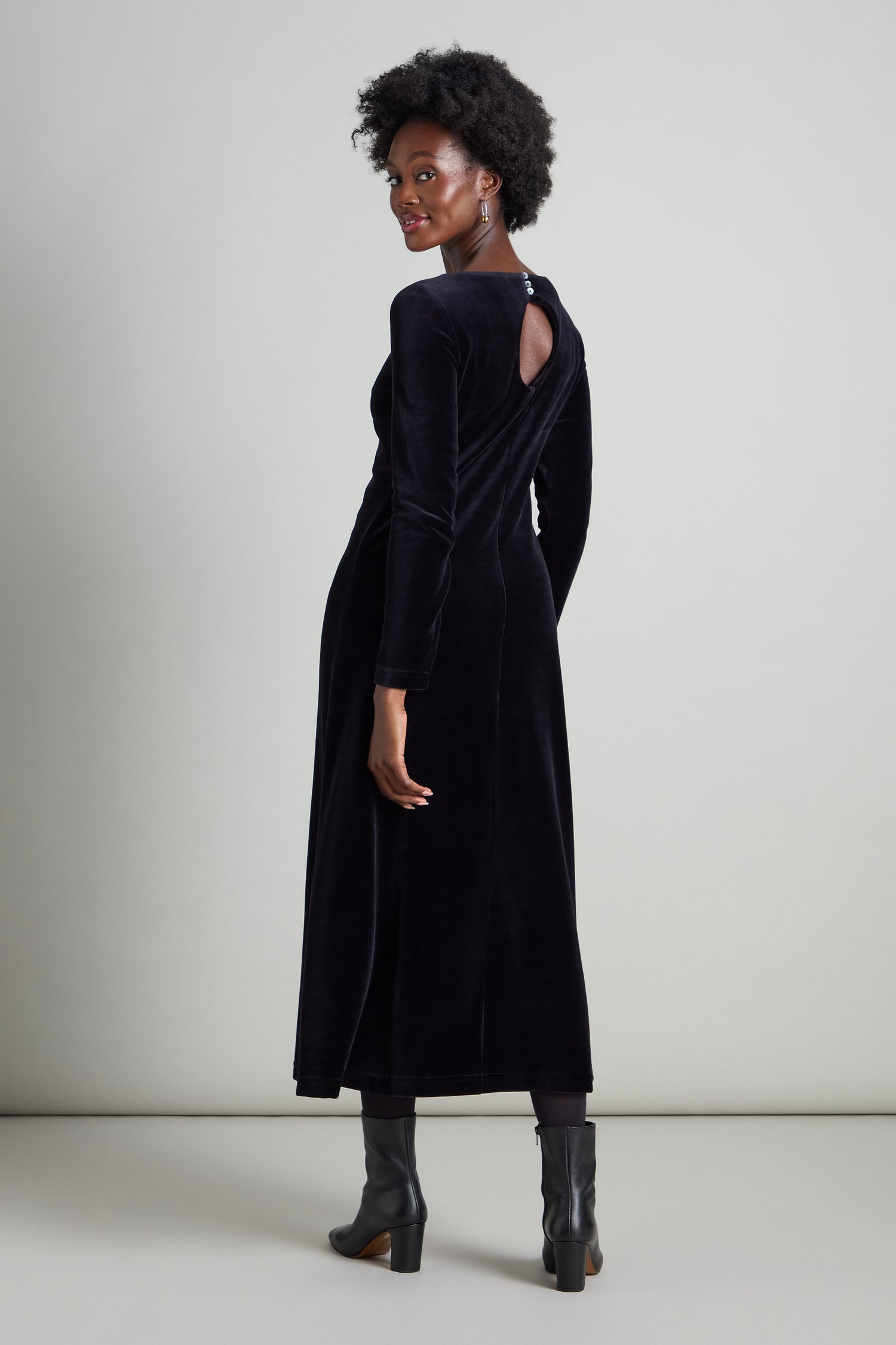 A woman in a Velvet Jersey Maxi A-Line Dress, featuring a fit-and-flare silhouette, stands gracefully against a neutral background, her black boots adding an air of understated glamour as she looks over her shoulder.