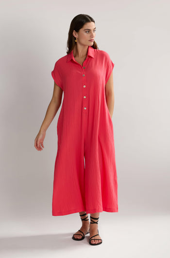 A person stands in a pink Crinkle Viscose Jumpsuit with short sleeves and button details, wearing black strappy sandals. The outfit is accented with an SPD5896-CSV tag, adding a touch of modernity to the classic look.