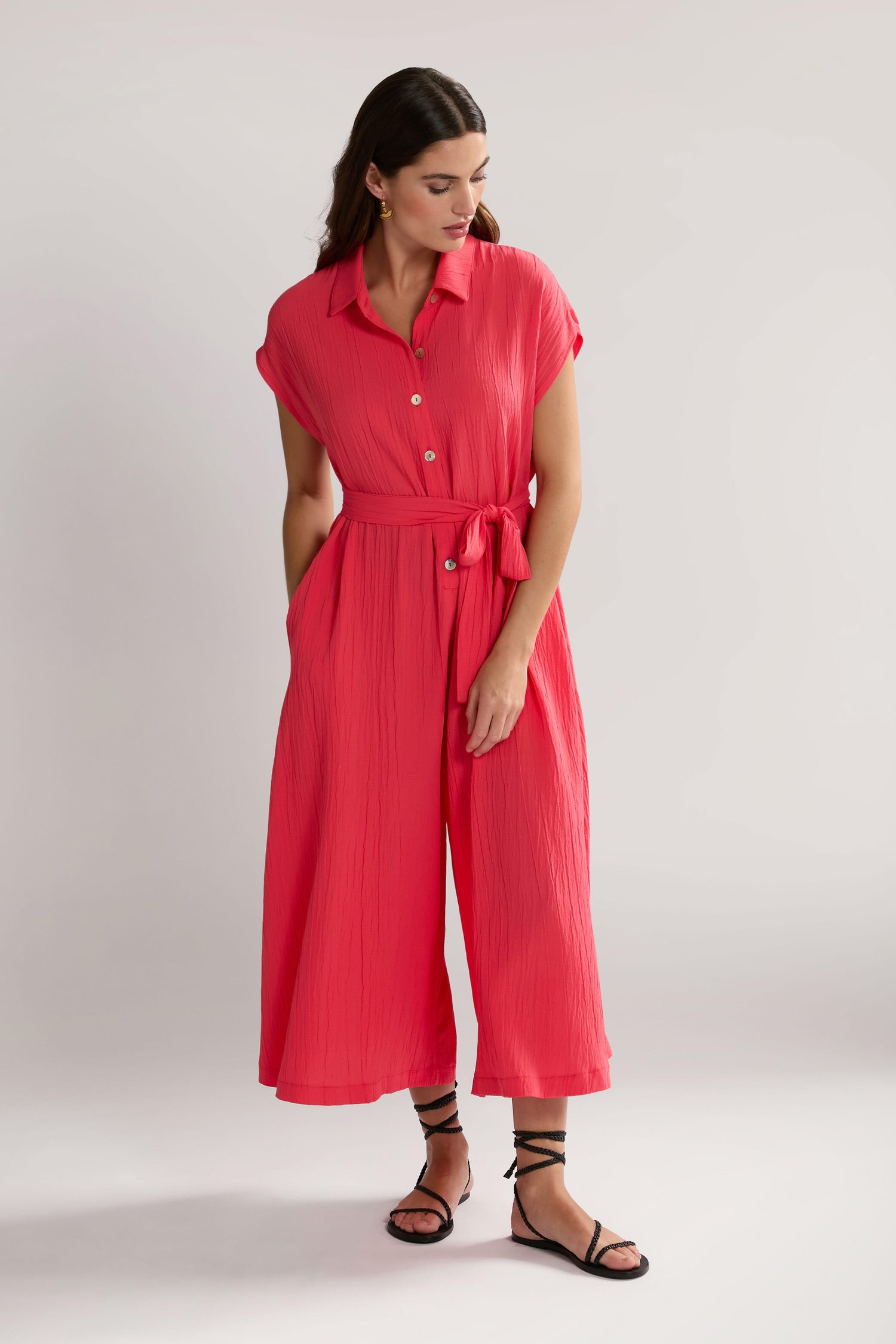 A person in a bright pink Crinkle Viscose Jumpsuit with a tied waist and black strappy sandals stands against a plain background, looking down with hands in pockets.