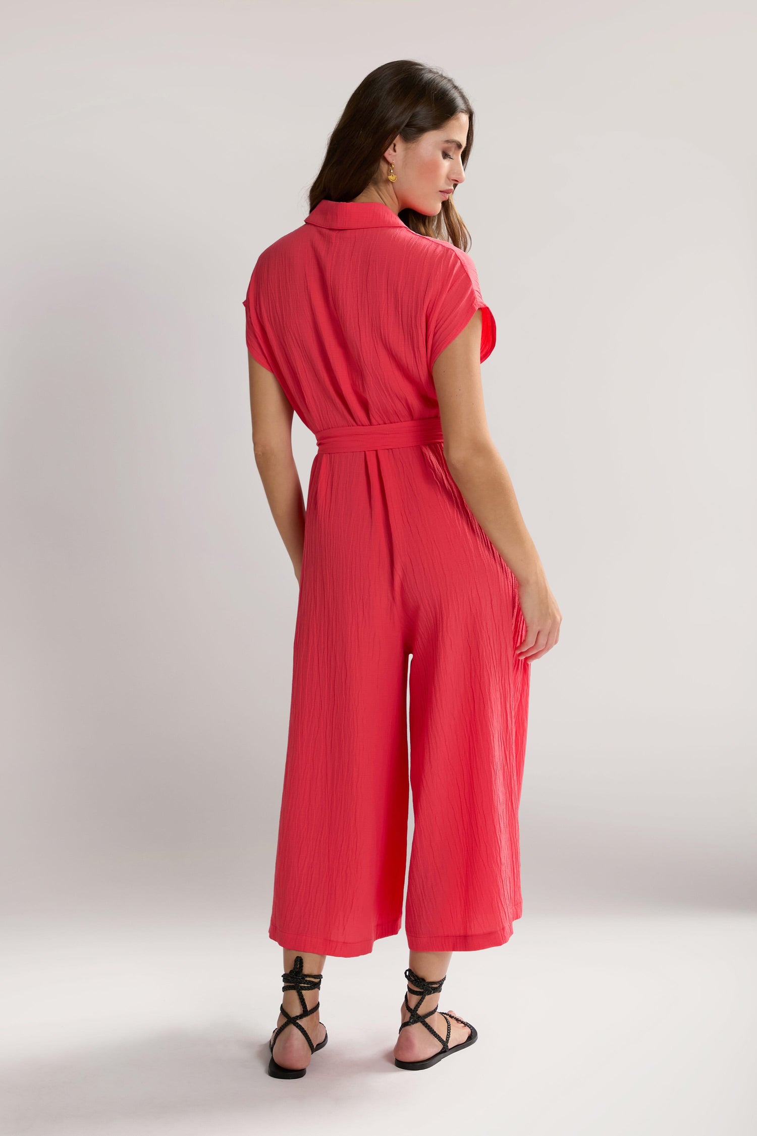 A woman stands facing slightly to the side, wearing a bright red, short-sleeved Crinkle Viscose Jumpsuit with a belt and black strappy sandals. The background is plain and light grey.