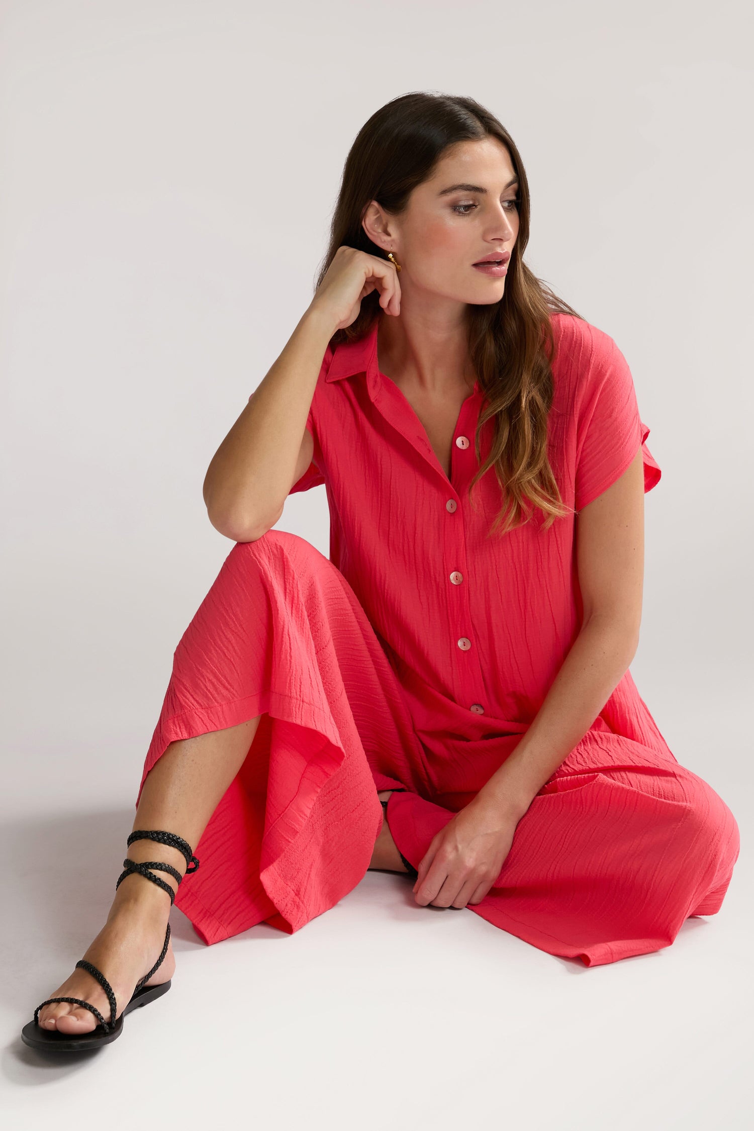 A woman in a red dress, labeled Crinkle Viscose Jumpsuit, sits on the floor with one leg crossed beneath her and the other bent. She looks to her right and rests her left elbow on her knee.