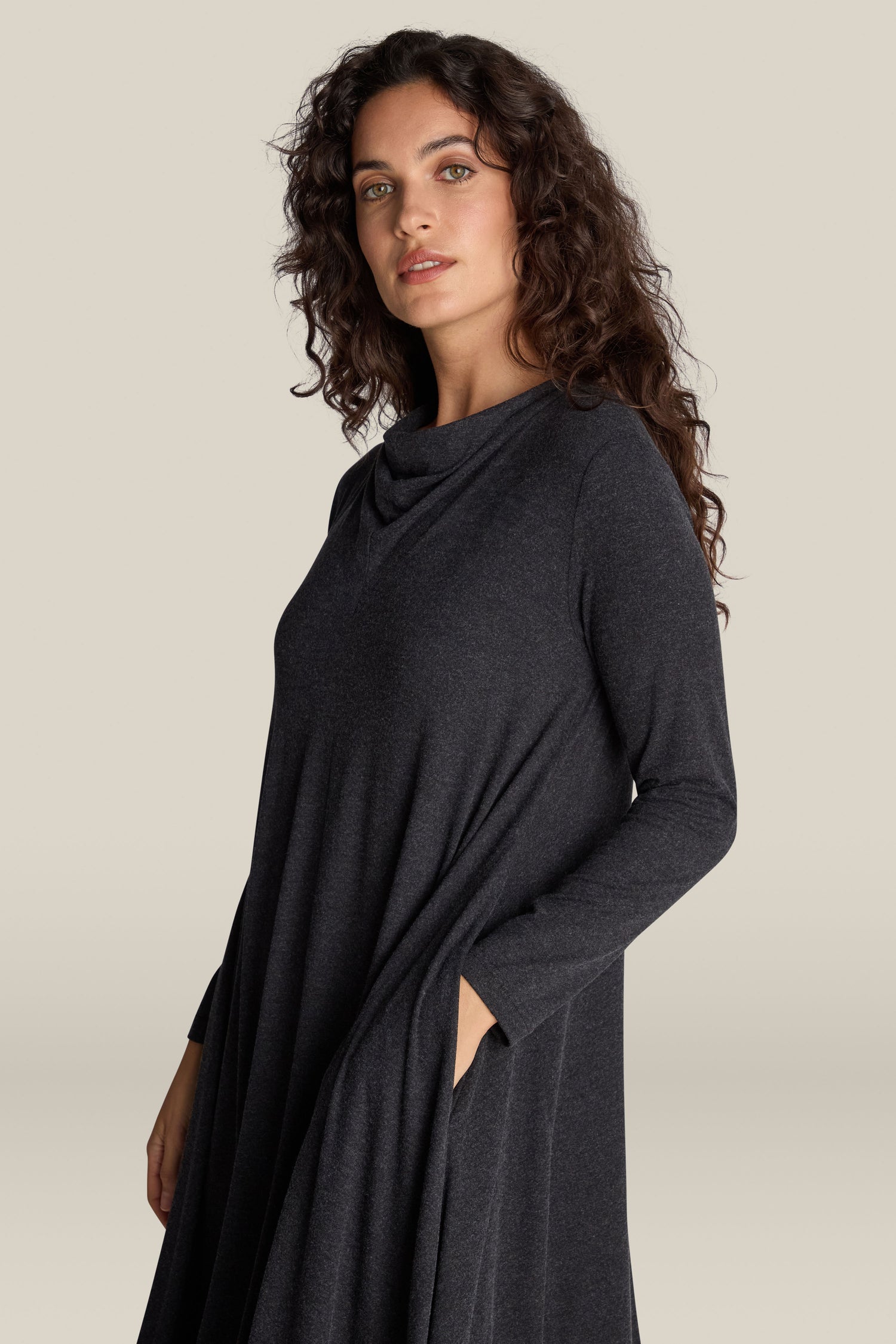 A person with long, wavy dark hair wears a Soft Viscose Jersey Flared Dress in dark gray, featuring loose, long sleeves and a draped neckline, standing against a plain background.