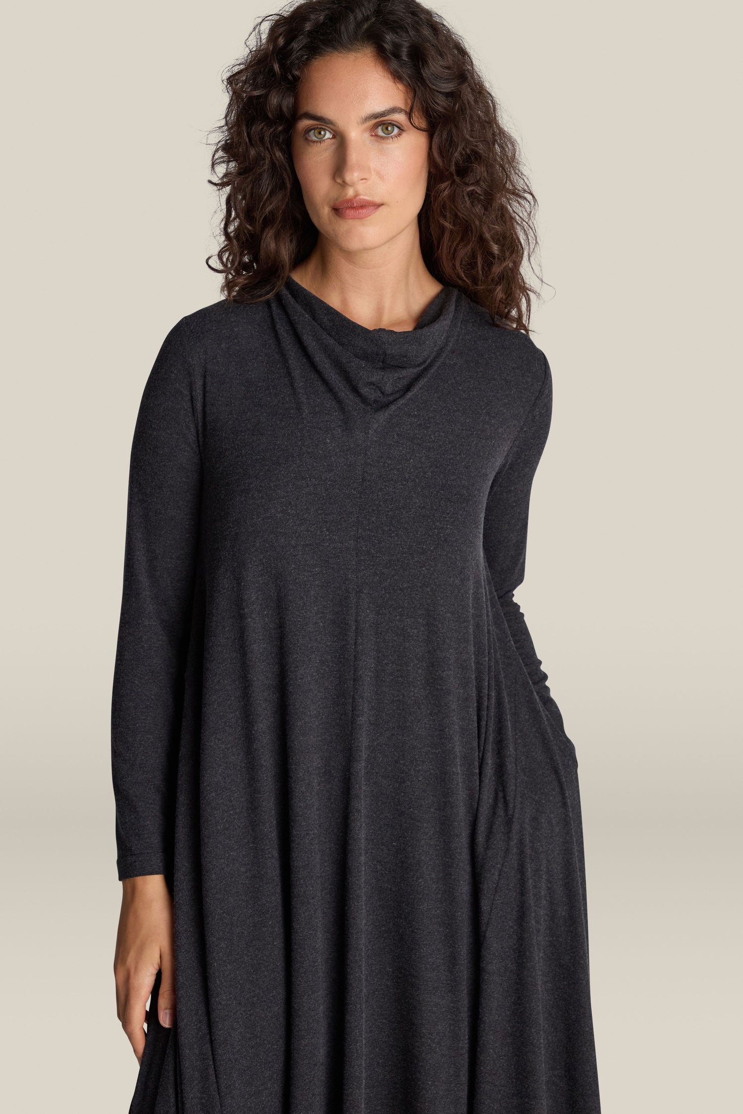 A woman with curly hair is wearing the Soft Viscose Jersey Flared Dress. The long-sleeve, dark-colored A-line silhouette contrasts elegantly against the plain, light background.