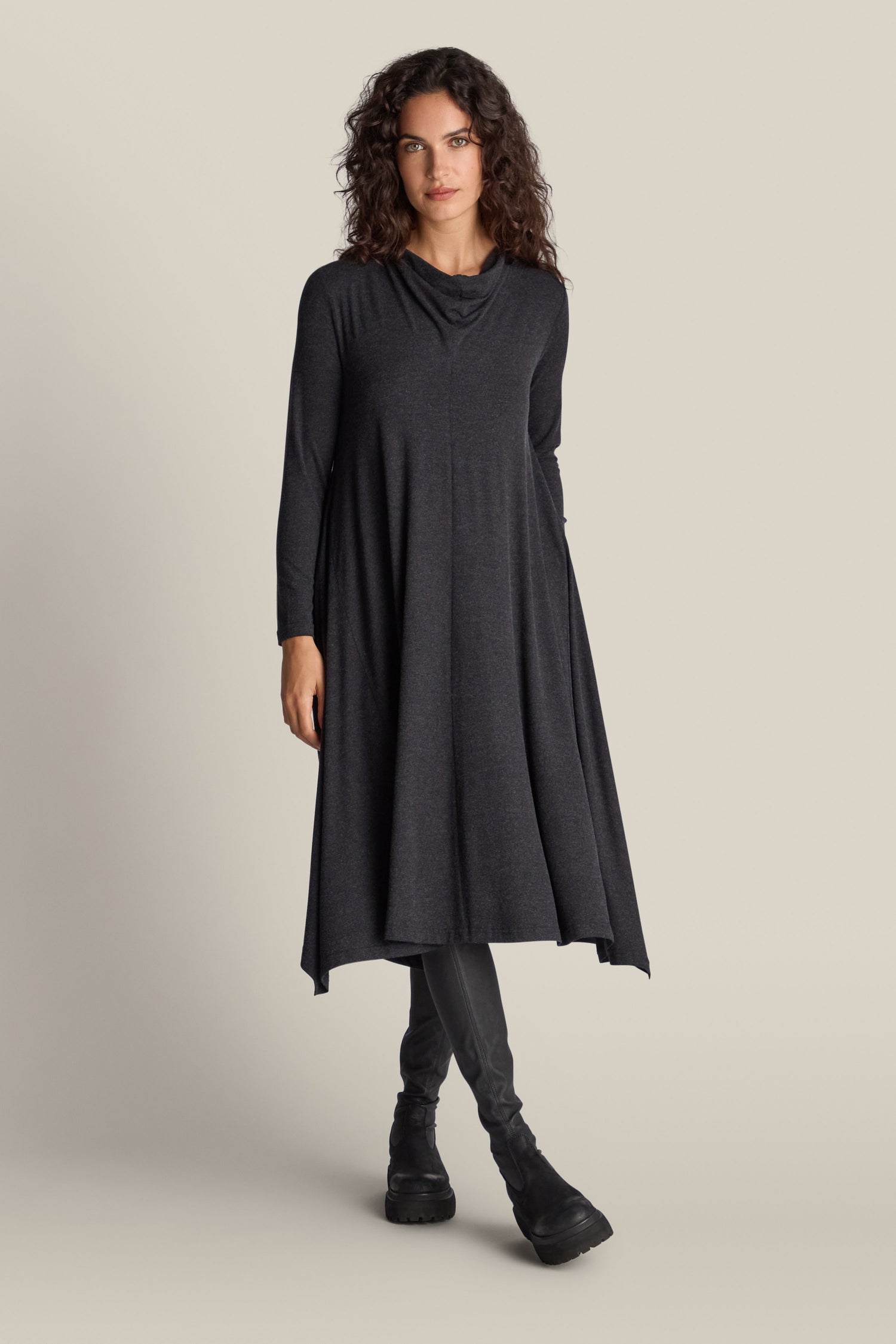 A woman stands against a plain background wearing the Soft Viscose Jersey Flared Dress in dark gray, featuring long sleeves and paired with black leggings. She has curly hair and complements her outfit with black boots, effortlessly highlighting the softness of the dress.