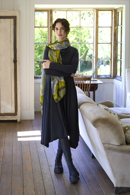 A woman stands in a well-lit living room wearing a Soft Viscose Jersey Flared Dress with an A-line silhouette, leggings, boots, and a patterned scarf. She has short curly hair and is positioned in front of a large window.