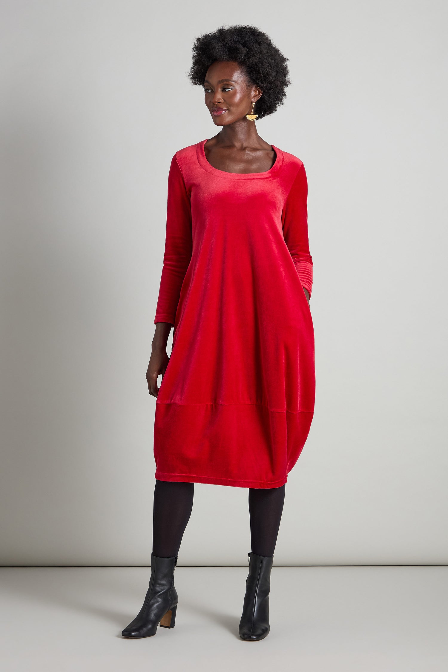 Dressed in a Velvet Jersey Bubble Dress featuring a horseshoe neckline and long sleeves crafted from plush, stretchy velvet fabric, an individual stands against a neutral background wearing black tights and ankle boots.