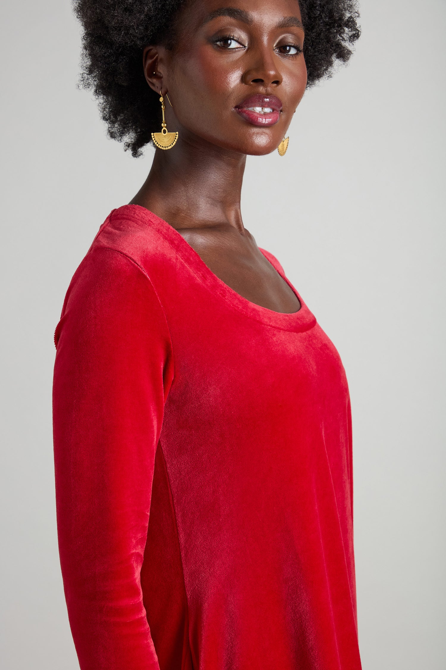 A person with curly hair is wearing a Velvet Jersey Bubble Dress in red, made from plush, stretchy velvet fabric, along with gold earrings, while looking at the camera against a plain background.