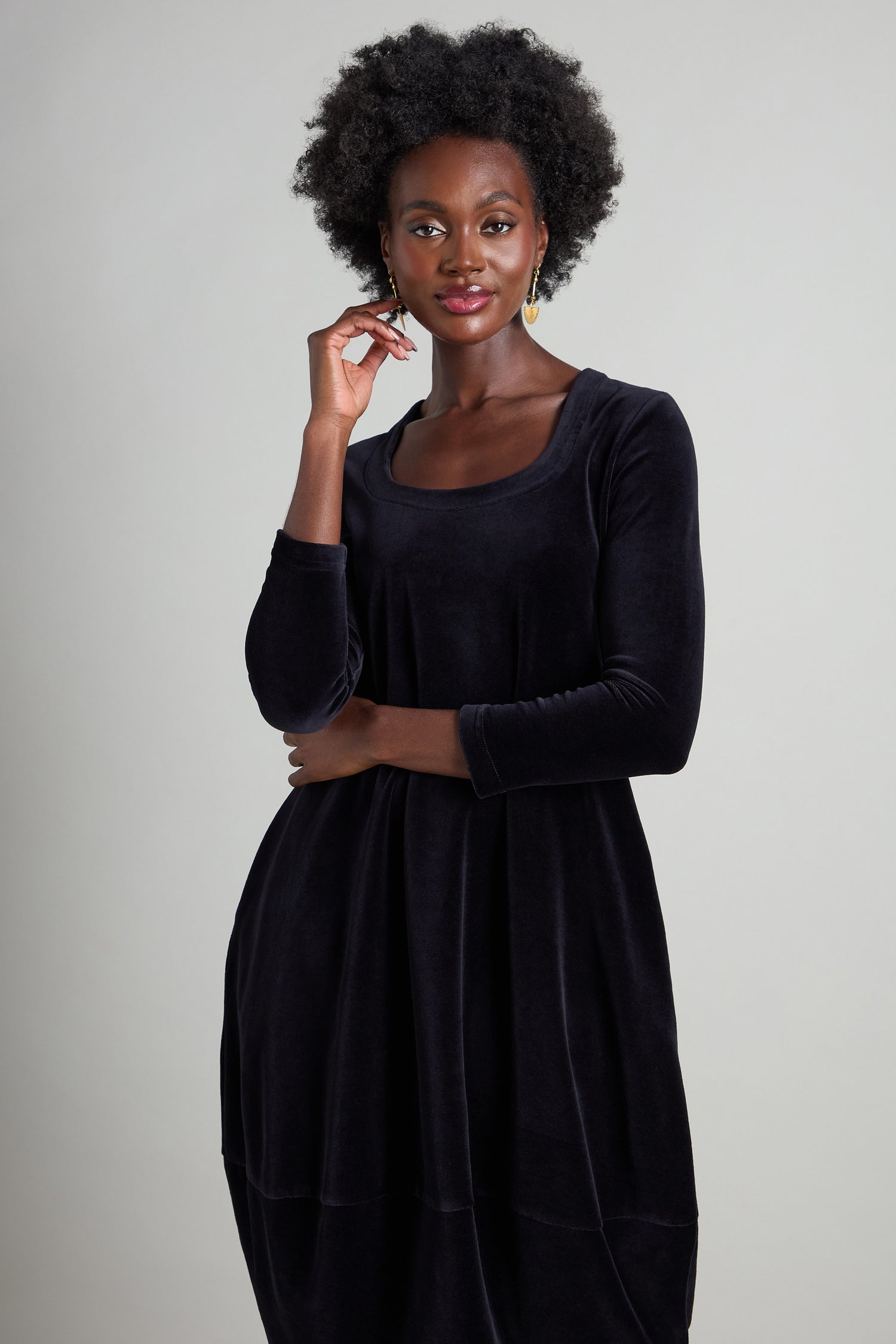 A person gracefully poses against a neutral background, wearing the Velvet Jersey Bubble Dress featuring long sleeves and a horseshoe neckline, gently touching their face with one hand.