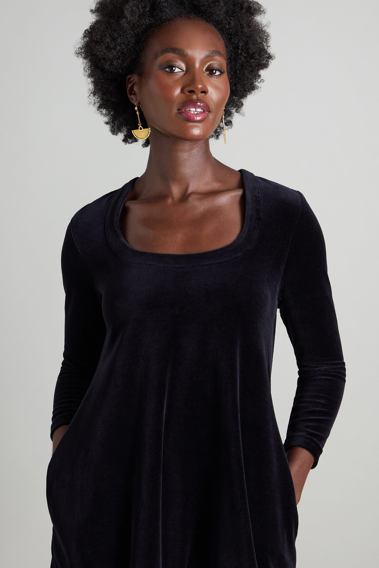 A person with curly hair poses against a neutral background, showcasing the Velvet Jersey Bubble Dress in black, known for its chic horseshoe neckline.