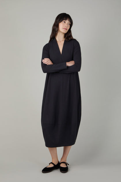 A woman stands with arms crossed, wearing the Fluid Crepe V Neck Black Bubble Dress—a loose, long-sleeve black crepe jersey piece with a subtle bubble silhouette—paired with black flats against a plain light gray background.