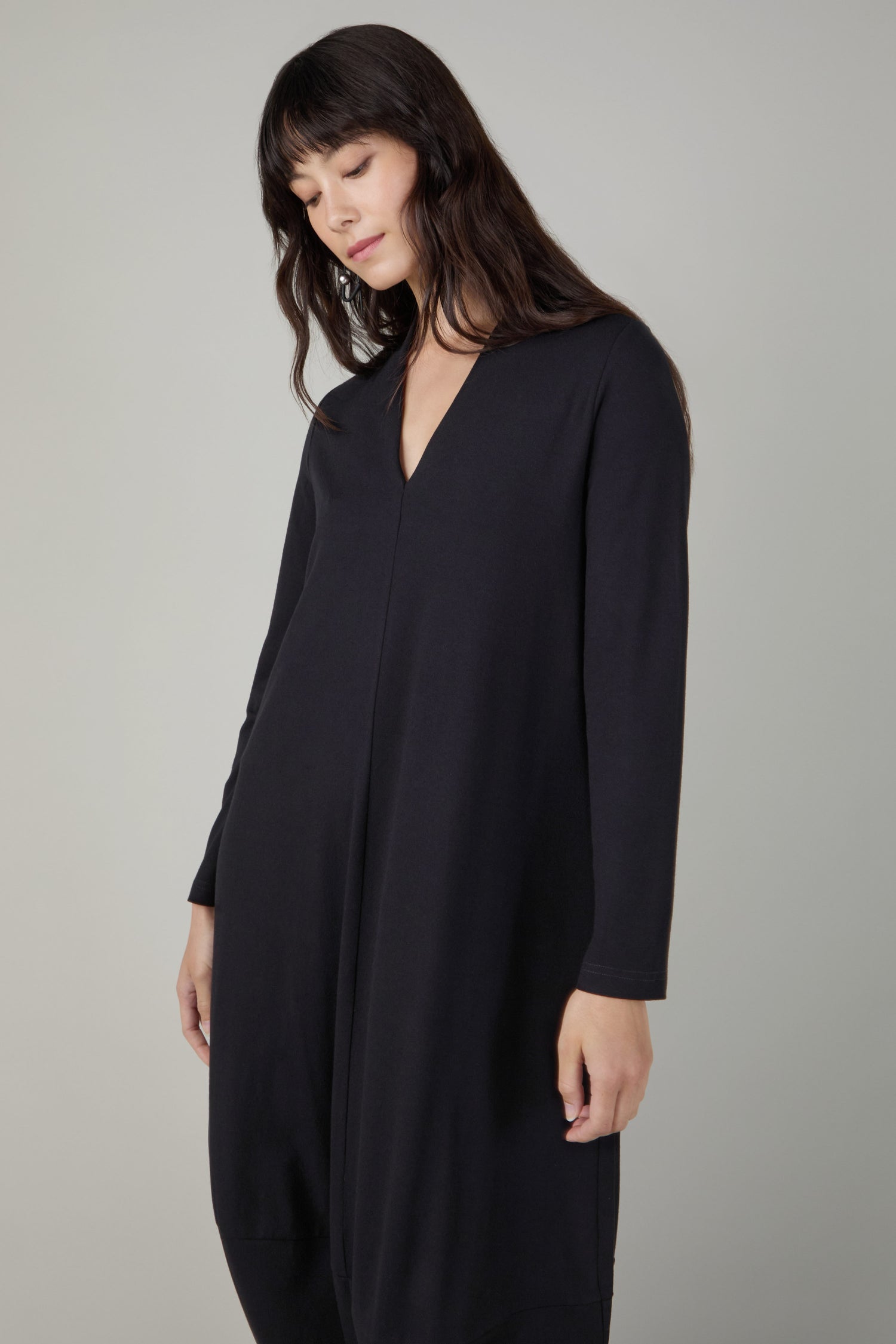 A woman with long dark hair looks down while wearing the Fluid Crepe V Neck Black Bubble Dress against a plain light gray background.