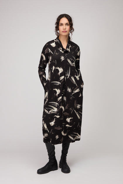 Woman wearing the Painted Brushstrokes Jersey Bubble Dress—black, long-sleeve, with white hand-painted brushstrokes and pockets—paired with black boots, stands against a plain light background.