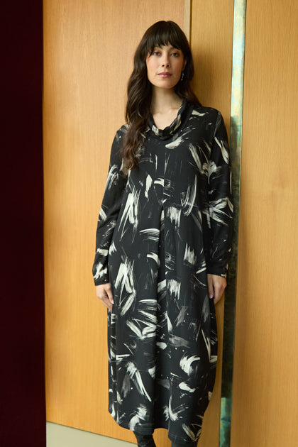 A woman stands indoors against a wooden wall, wearing the Painted Brushstrokes Jersey Bubble Dress—a long-sleeved black viscose jersey dress featuring white abstract brushstroke patterns.