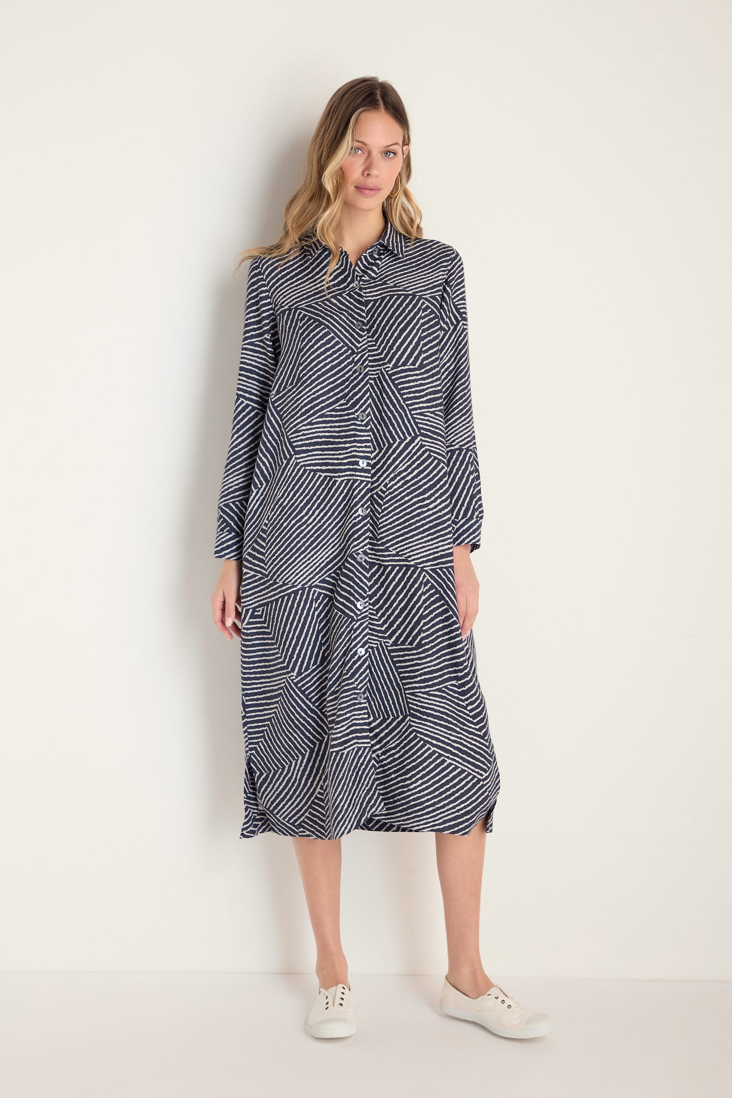 A woman models the Linear Prism Shirt Dress, featuring long sleeves and a navy blue and white geometric print in brushed viscose, styled with white sneakers against a plain light background.