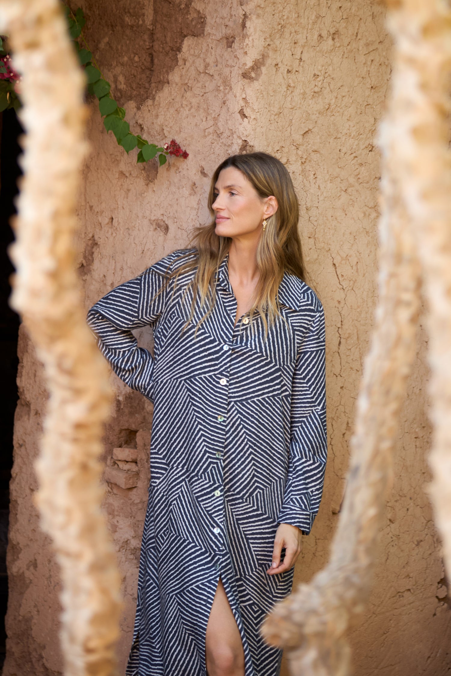 A woman wearing the Linear Prism Shirt Dress with a geometric print stands before a textured brown wall, framed by two blurred tree branches.