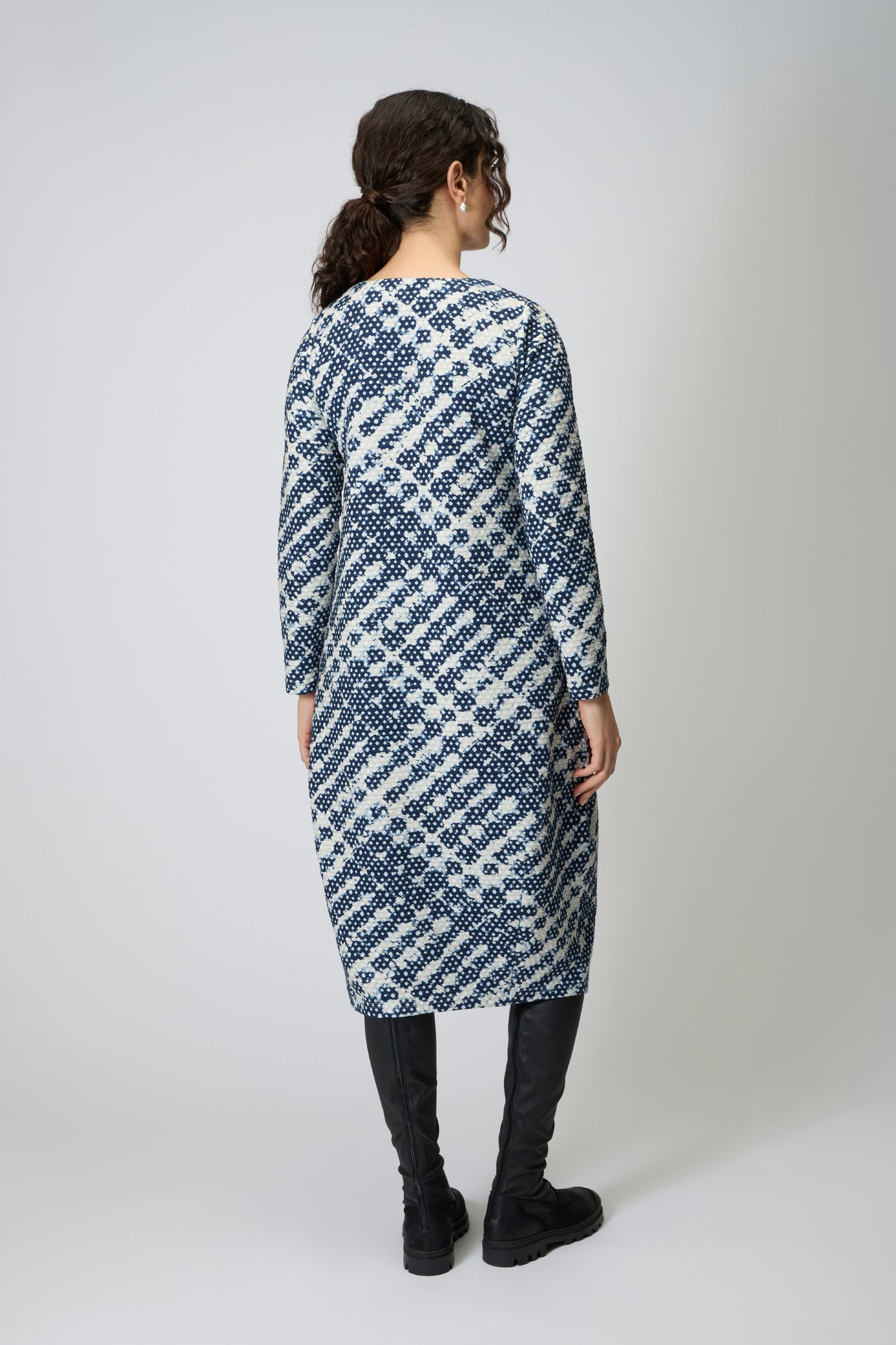 A person faces away wearing the Diamond Tie Dye Jersey Bubble Dress— a long-sleeved, knee-length, bubble silhouette dress in navy and white viscose jersey—paired with black knee-high boots.