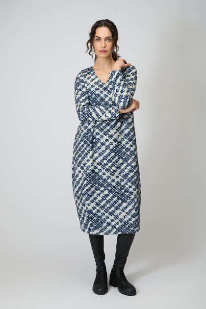 A woman with curly hair and a neutral expression stands against a plain background wearing the Diamond Tie Dye Jersey Bubble Dress, styled with a belt, black leggings, and boots.