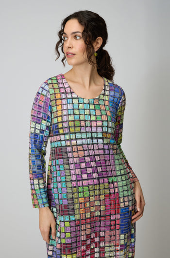 A woman wearing the Coloured Glass Tiles Jersey Bubble Dress, featuring vibrant long sleeves and a mosaic-inspired square pattern, looks to her left against a plain background.